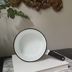 May include: A white enamel saucepan with a black rim and handle. The pan sits on a white book atop a marble surface. A gold ornate mirror is in the background.