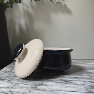 Könnte beinhalten: Ein kleiner, runder, marineblauer Keramikbehälter mit passendem Deckel. Der Deckel ist cremefarben mit einem marineblauen Knopf. Der Behälter steht auf einer grau-weißen Marmoroberfläche. Im Hintergrund befindet sich eine schwarze Vase mit Grün.
