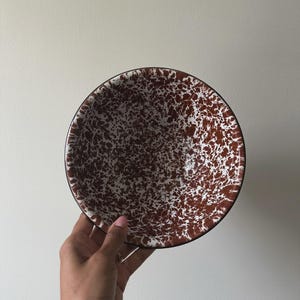May include: A round, speckled bowl with a brown and white pattern. The bowl has a black rim and is held by a hand against a white background. The bowl's design is reminiscent of enamelware.
