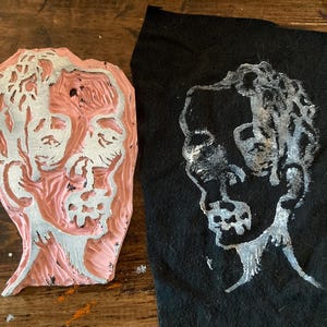 May include: A pink rubber stamp with a detailed carving of a face. The stamp is next to a black fabric with a white print of the same face.