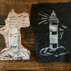 May include: A lighthouse block print and its impression on black fabric. The block is a light pink and gray color, with a detailed lighthouse design. The fabric impression is a white lighthouse on a black background.