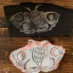 May include: A close-up of a wooden butterfly stamp with a pink base and a silver butterfly design. The stamp is used to create a print on fabric.