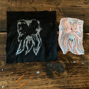 May include: A close-up of a black fabric with a white design of two figures with long hair. Next to the fabric is a pink rubber stamp with a similar design.