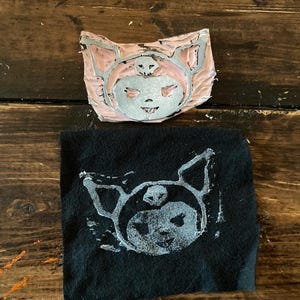 May include: A close-up of a black fabric with a white print of a cartoon character with a skull and crossbones. The character is wearing a pink hat. The print is from a carved rubber stamp.
