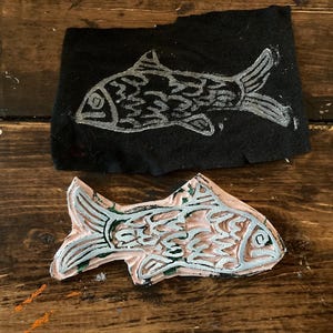 May include: A close-up of a carved wooden fish stamp. The stamp is painted pink and has a detailed design of a fish with scales. The stamp is on a wooden surface.