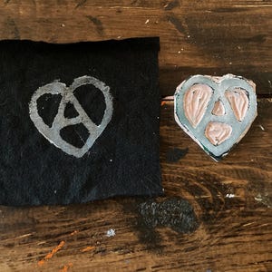 May include: A white peace sign inside a pink heart shape, printed on a black fabric. The heart shape is also a separate object, made of a textured material.
