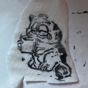 May include: Black and white ink drawing of a cartoon character with large eyes and a hat, printed on a white fabric patch.