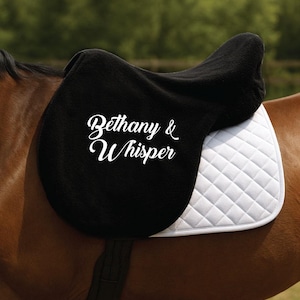 Può includere: Un sottosella nero per cavalli con la scritta "Bethany & Whisper" in corsivo bianco. Il sottosella è su un cavallo marrone, con un sottosella trapuntato bianco sotto.