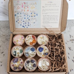 May include: A gift box containing nine small candles in copper-colored tins. Each candle is topped with crystals and dried botanicals. The box also includes a card with text and a list of candle scents.