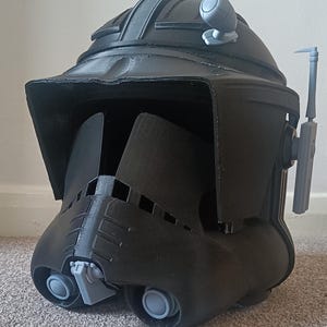 May include: A black helmet with gray accents, featuring a visor, a top ridge, and side attachments. The helmet has a rounded shape with detailed features, including vents and a chin piece. The design suggests a futuristic or sci-fi theme.