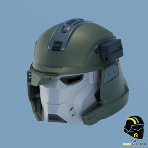 Könnte beinhalten: Ein futuristischer Helm in Olivgrün und Grau mit weißem Gesichtsschutz. Der Helm hat eckige Linien, ein Visier und ein oben angebrachtes Detail. Das Prop Spector-Logo befindet sich in der unteren rechten Ecke.