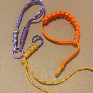 May include: Three handmade paracord accessories: a purple and white lanyard with a lavender ring, an orange bracelet, and a yellow and blue keychain with a blue ring. The items are made of woven nylon cord.