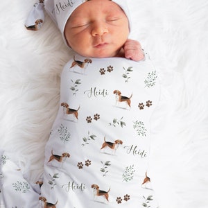 May include: A white swaddle with a pattern of brown and white beagle dogs, green leaves, and brown paw prints. The swaddle has the name "Heidi" printed on it. A white hat with the same pattern is on the baby's head.