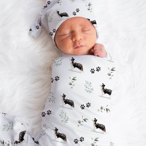 May include: A newborn baby swaddled in a white swaddle blanket with a black and white border collie print. The baby is wearing a matching hat with the same print. The blanket and hat are decorated with small green leaves and black paw prints.
