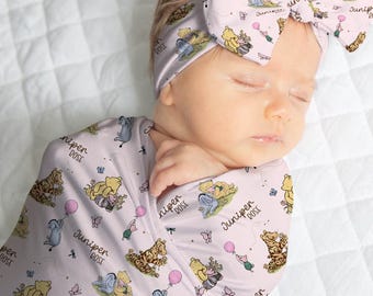May include: A light pink swaddle blanket and matching headband set featuring a repeating pattern of cartoon characters, including a yellow bear, a gray donkey, and a small pig, along with pink balloons and the text Juniper Rose.