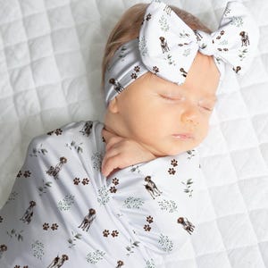 May include: A white swaddle with a brown and white dog print and a matching headband with a bow. The baby is sleeping peacefully.