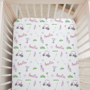 May include: White crib sheet with a pink and green golf-themed pattern. The sheet features golf carts, golf clubs, golf balls, and the name "Amelia" repeated throughout.