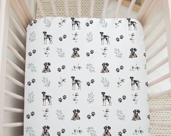 May include: A white fitted crib sheet with a repeating pattern of black and gray dogs, paw prints, and green leaves. The sheet is inside a light pink wooden crib.