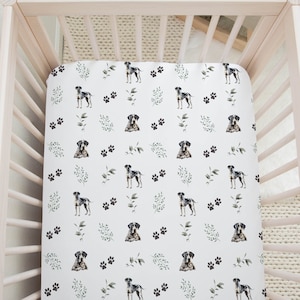 May include: A white fitted crib sheet with a repeating pattern of black and gray dogs, paw prints, and green leaves. The sheet is inside a light pink wooden crib.