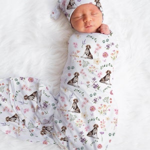 May include: A newborn baby swaddled in a white blanket and matching hat, both adorned with a floral and dog print. The design features watercolor-style German Shorthaired Pointers, pink and purple flowers, and green paw prints.