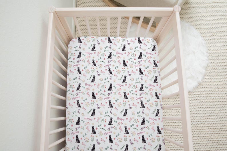 May include: A white fitted crib sheet with a repeating pattern of black dogs and pink flowers. The sheet has the name "Harper" printed on it.