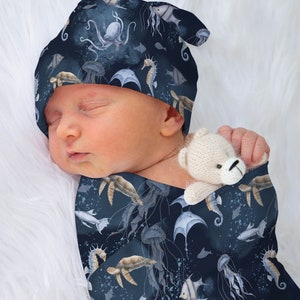 May include: A baby wearing a blue and white swaddle with a sea life pattern. The swaddle features seahorses, jellyfish, turtles, and fish. The baby is also wearing a matching blue and white hat with the same sea life pattern.