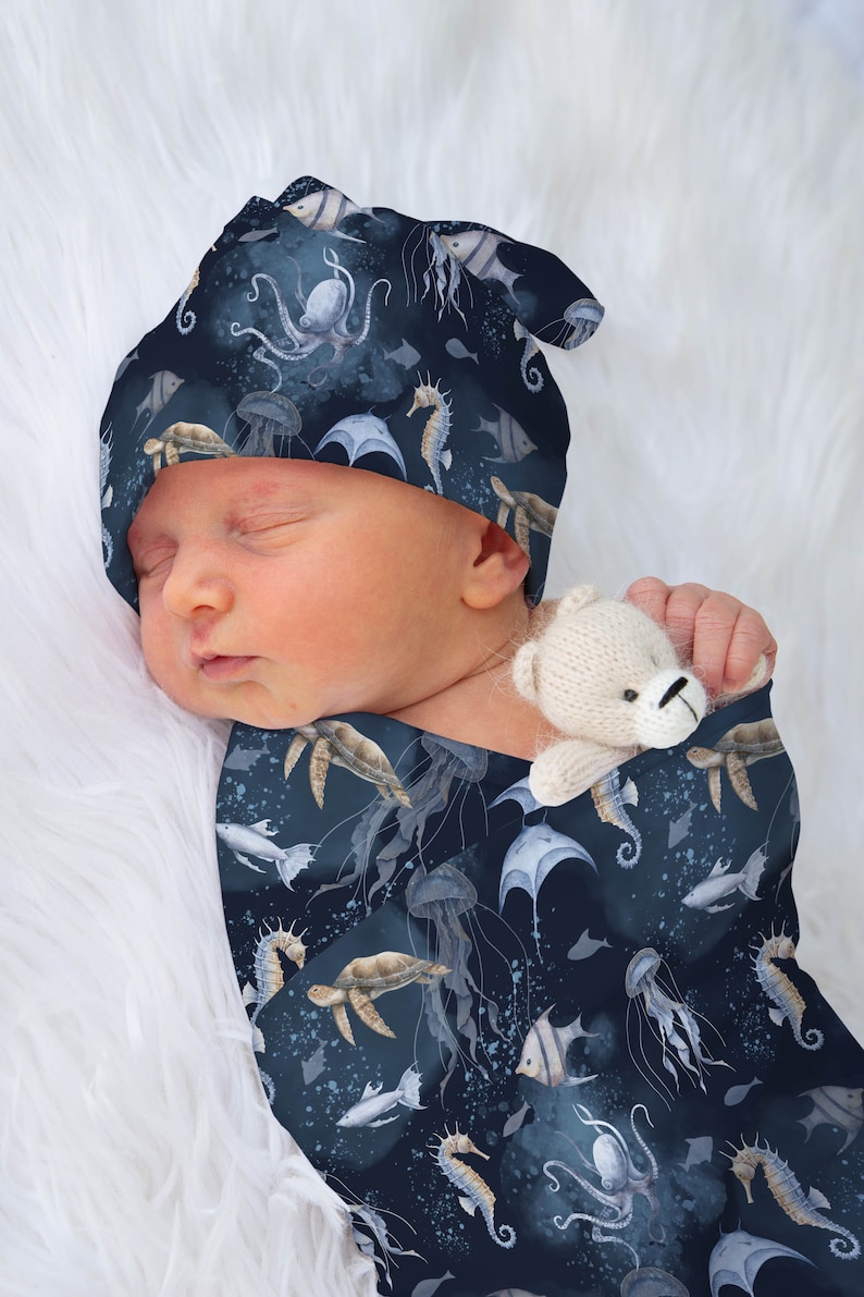 May include: A baby wearing a blue and white swaddle with a sea life pattern. The swaddle features seahorses, jellyfish, turtles, and fish. The baby is also wearing a matching blue and white hat with the same sea life pattern.