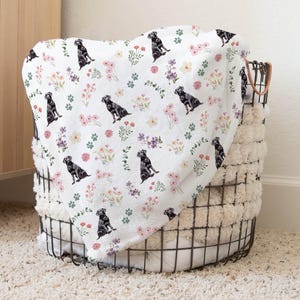 May include: A white throw blanket with a black Labrador Retriever and floral pattern. The blanket is folded and placed in a wire basket.