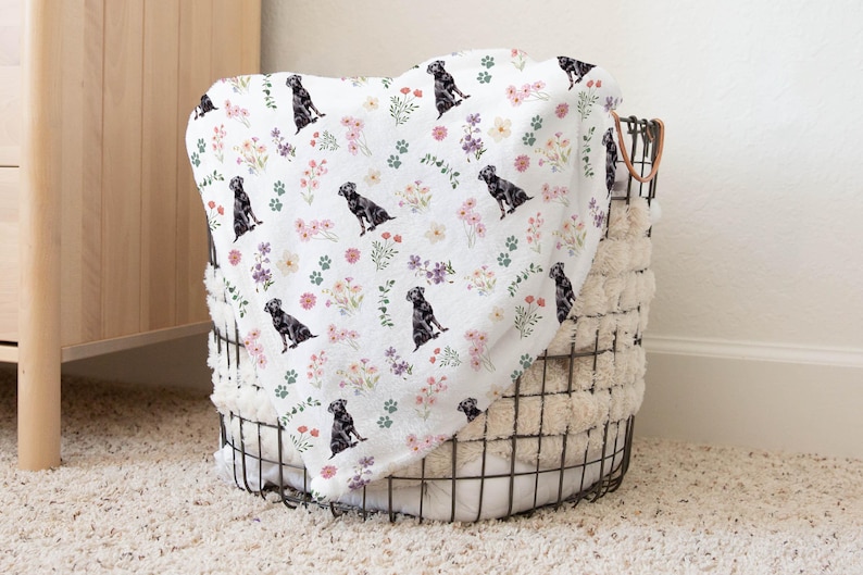 May include: A white throw blanket with a black Labrador Retriever and floral pattern. The blanket is folded and placed in a wire basket.