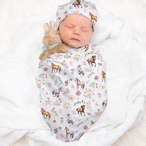 May include: A newborn baby swaddled in a white and pink swaddle with a horse and floral print. The baby is wearing a matching hat with a horse and floral print. The baby is holding a stuffed bunny rabbit.