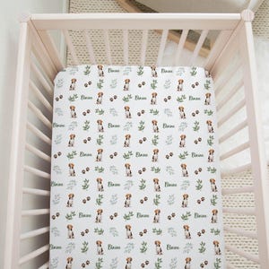 May include: White crib sheet with a repeating pattern of brown and white dogs, green leaves, and the name "Blaine" printed in green.