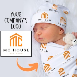 May include: A white swaddle blanket with a repeating pattern of the logo "MC House Building". A baby is wearing a matching hat with the same logo. The baby is sleeping and holding a white teddy bear.