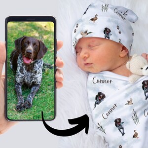 May include: A white swaddle blanket with a repeating pattern of brown and white dogs, ducks, and reeds. The blanket has the name "Connor" printed on it multiple times. A baby is sleeping on the blanket, wearing a white hat with the same pattern as the blanket.