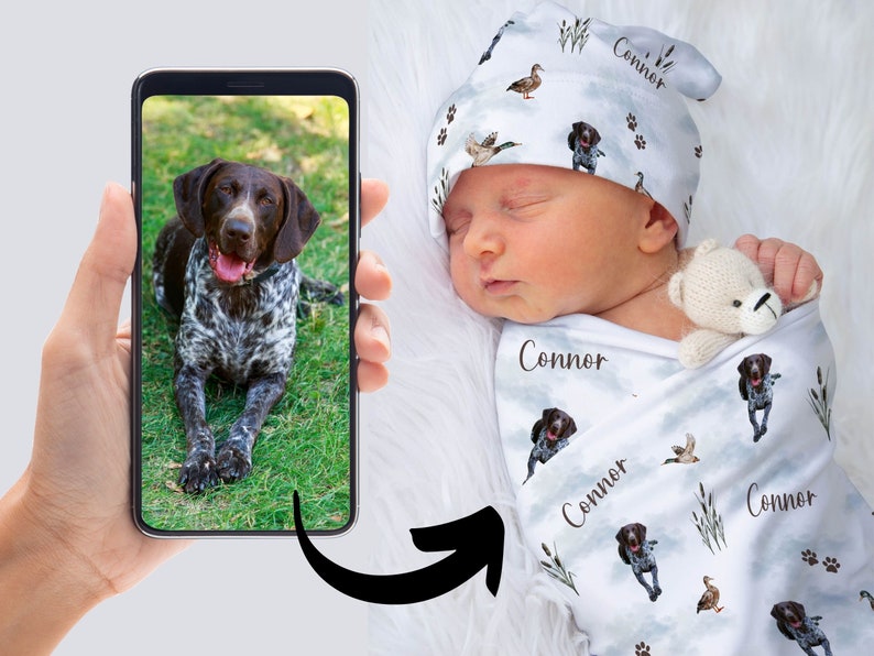 May include: A white swaddle blanket with a repeating pattern of brown and white dogs, ducks, and reeds. The blanket has the name "Connor" printed on it multiple times. A baby is sleeping on the blanket, wearing a white hat with the same pattern as the blanket.