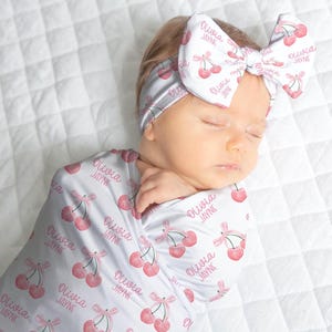 May include: A newborn baby wrapped in a white swaddle blanket and wearing a matching headband, both adorned with a cherry pattern and the name "Olivia Jayne" in pink. The baby is lying on a white quilted surface.