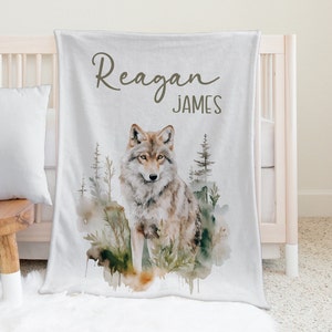 May include: A white baby blanket with a watercolor illustration of a wolf in a forest setting. The blanket has the name "Reagan James" printed on it in brown cursive.