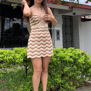 Puede incluir: Un vestido mini de crochet marrón y blanco con un patrón ondulado. El vestido tiene un diseño sin tirantes con un detalle anudado en la parte delantera. El vestido tiene un recorte en la parte delantera. El vestido lo lleva una persona de pie en una acera frente a una casa.