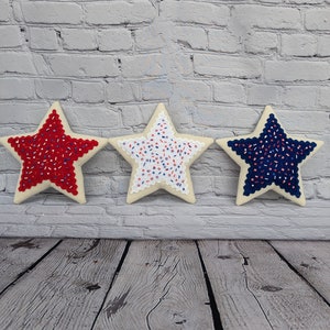 May include: Three felt star-shaped pillows with red, white, and blue sprinkles. The stars are arranged in a row on a wood floor in front of a white brick wall.