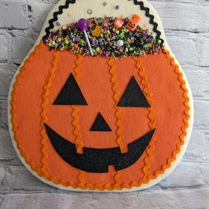May include: A felt pumpkin door hanger with a smiling jack-o'-lantern face. The pumpkin is filled with colorful sprinkles, candy, and a lollipop.