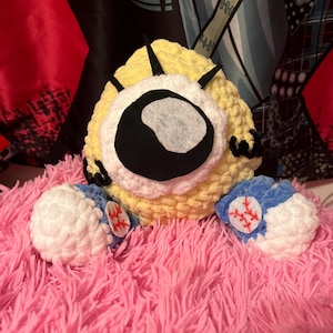 Könnte beinhalten: Ein gehäkeltes Plüschtier in Form einer Comicfigur mit einem einzigen großen Auge, gelbem Körper und blauen und weißen Armen mit Baseballnähten. Das Spielzeug sitzt auf einer rosa flauschigen Oberfläche.