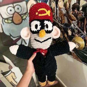 Könnte beinhalten: Gehäkeltes Plüschtier einer Figur aus der Zeichentrickserie Gravity Falls. Das Spielzeug hat einen roten Hut mit gelbem Emblem, eine schwarze Brille, ein weißes Gesicht und einen schwarzen Anzug. Das Spielzeug wird von einer Hand gehalten.