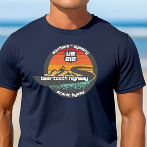 以下が含まれることがあります： 「Montana - Wyoming US 212 Beartooth Highway Scenic Byway」というテキストが書かれた、山道が描かれたヴィンテージスタイルのグラフィックが施されたネイビーブルーのTシャツです。