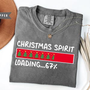 May include: A gray t-shirt with the words "CHRISTMAS SPIRIT LOADING...67%" in white. The design includes a red bar with Christmas trees. A silver necklace with a pendant is on the shirt.