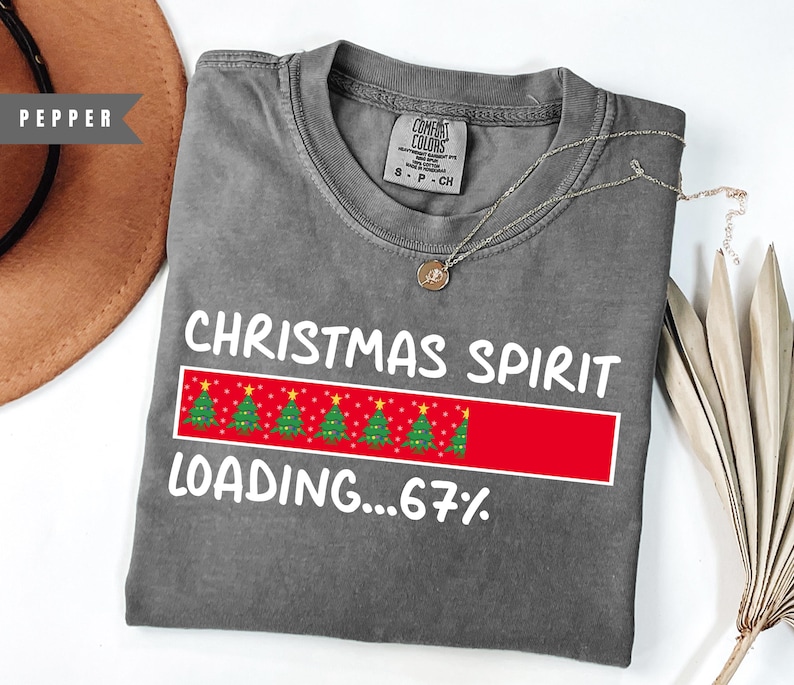 May include: A gray t-shirt with the words "CHRISTMAS SPIRIT LOADING...67%" in white. The design includes a red bar with Christmas trees. A silver necklace with a pendant is on the shirt.