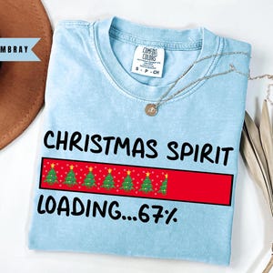 May include: Light blue t-shirt with the words "CHRISTMAS SPIRIT LOADING...67%" in black. The design includes a red rectangle with green Christmas trees. A silver necklace with a pendant is on the shirt.