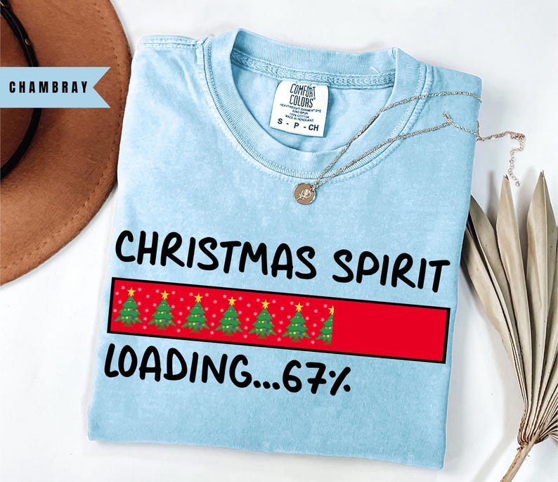 May include: Light blue t-shirt with the words "CHRISTMAS SPIRIT LOADING...67%" in black. The design includes a red rectangle with green Christmas trees. A silver necklace with a pendant is on the shirt.