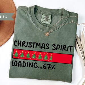 May include: A moss green t-shirt with the text "CHRISTMAS SPIRIT LOADING...67%" in black. The design includes a red loading bar with Christmas trees. A gold necklace is draped across the shirt.