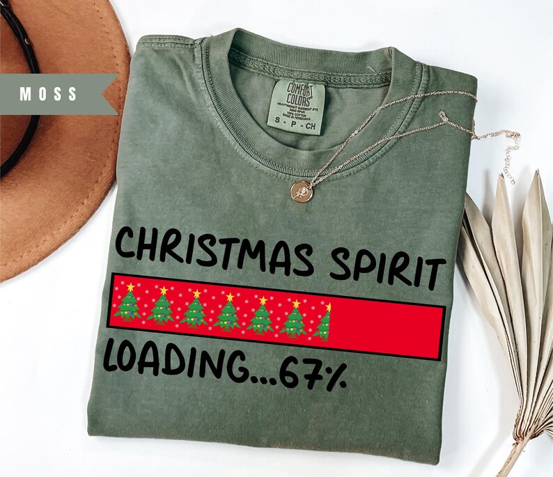 May include: A moss green t-shirt with the text "CHRISTMAS SPIRIT LOADING...67%" in black. The design includes a red loading bar with Christmas trees. A gold necklace is draped across the shirt.