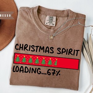 May include: A brown t-shirt with the words "CHRISTMAS SPIRIT" and "LOADING...67%" printed in black. A red bar with green Christmas trees is in the center. A silver necklace with a small pendant is on the shirt.