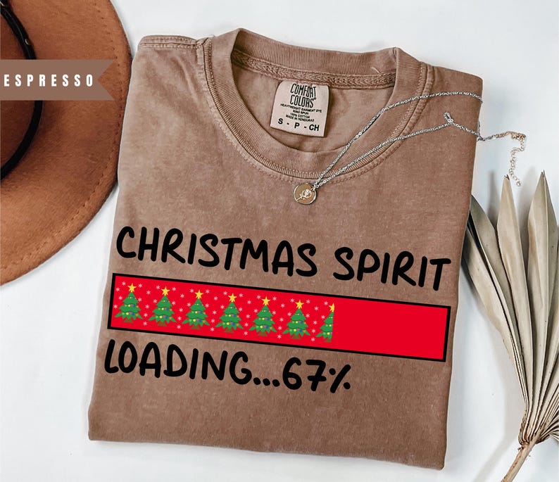 May include: A brown t-shirt with the words "CHRISTMAS SPIRIT" and "LOADING...67%" printed in black. A red bar with green Christmas trees is in the center. A silver necklace with a small pendant is on the shirt.